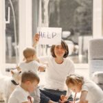 Mamá agotada en el suelo mientras cuida a sus hijos, sosteniendo un cartel que dice ayuda: el burnout de mamá cuidadora.